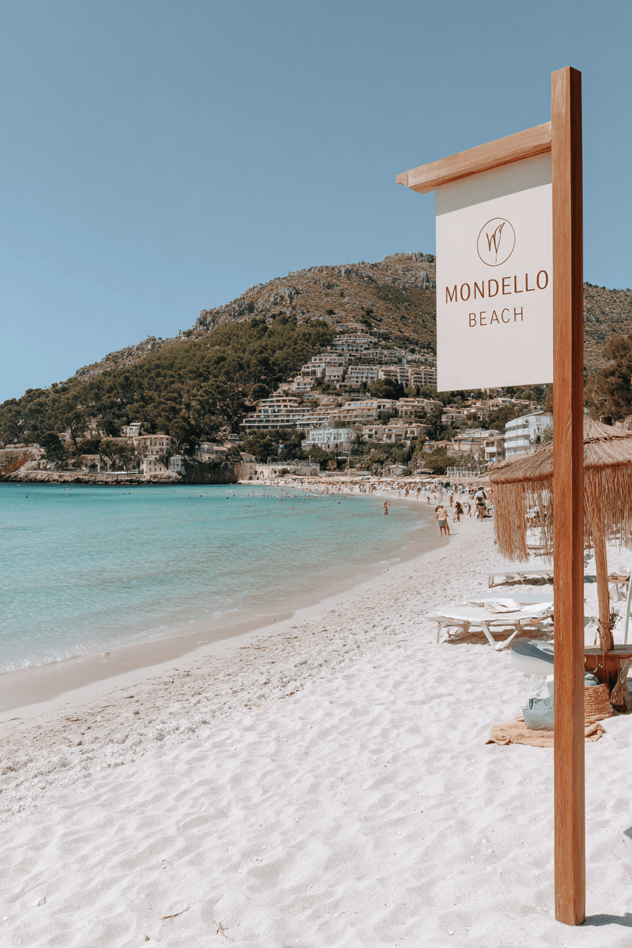 Schild von Mondello Beach am weißen Sandstrand mit türkisblauem Meer und Palermos Bergen im Hintergrund